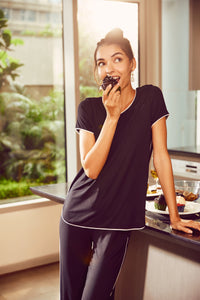 Black piping pajama set model enjoying a cupcake in a cozy kitchen.